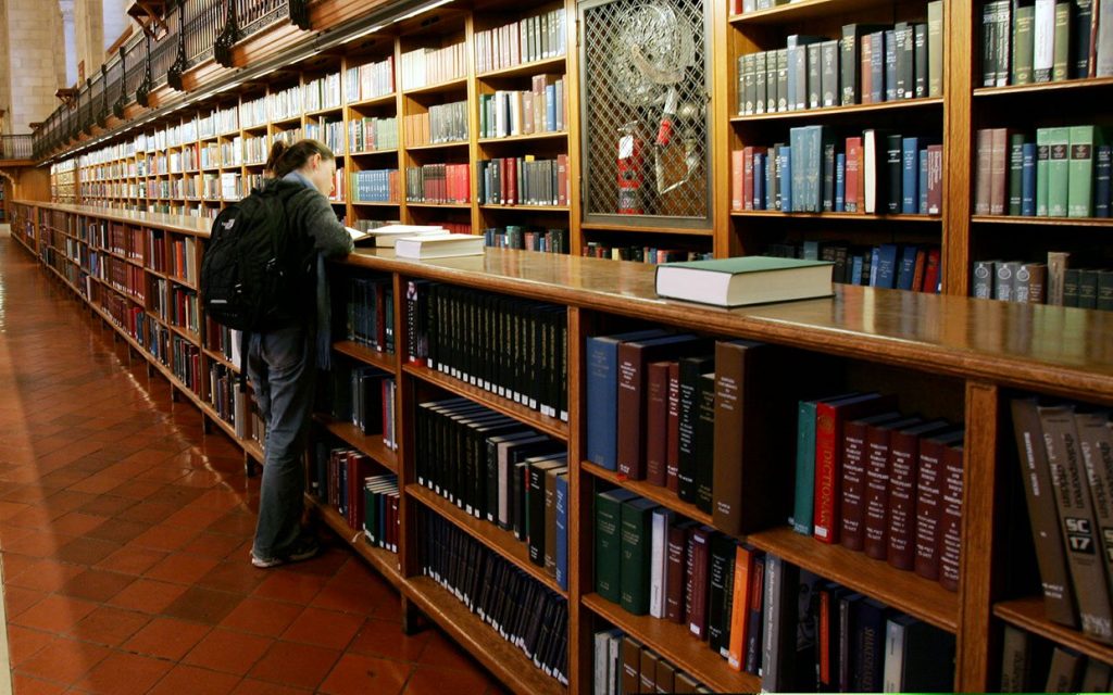 A library interior with long rows of wooden bookshelves filled with books and a person reading.